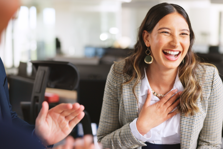 Two people in a modern office setting engaging in a conversation, with one person placing a hand on their chest while another gestures with their hands.